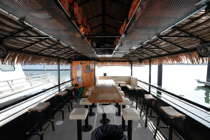 Boat interior with bar stools, wooden table, and thatched roof, docked by water.