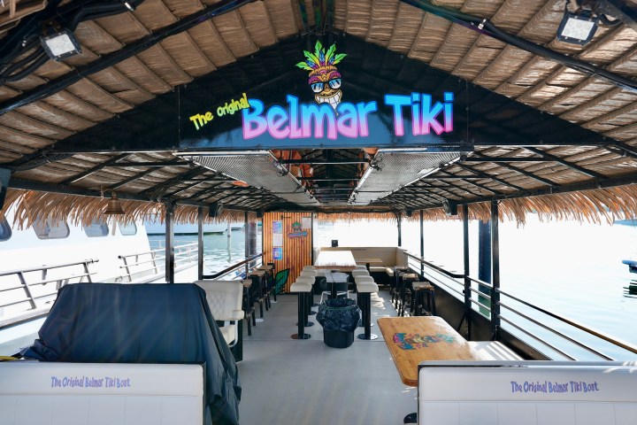 Interior of a thatched-roof boat with 'Belmar Tiki' sign, seating, and tables over water.