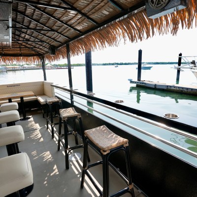 Tiki bar with stools and thatched roof on a boat docked by water in sunny weather.