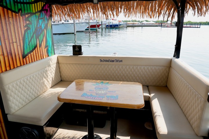 Interior of a Tiki boat with cushioned seating and a wooden table, overlooking a marina.