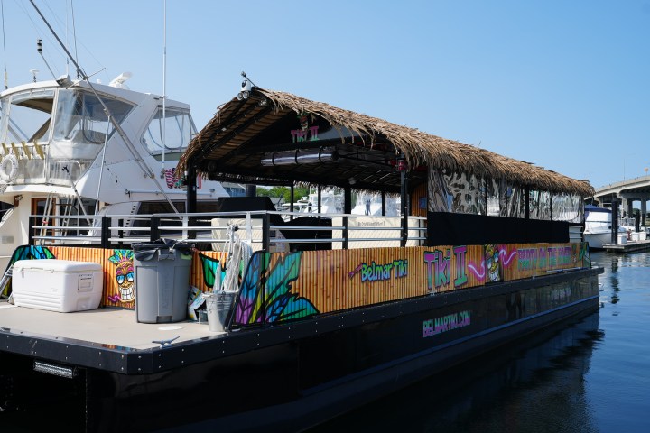 A tiki-themed boat with a thatched roof is docked at a marina on a sunny day.