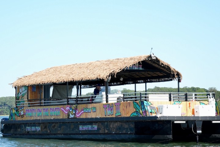 A tiki-themed pontoon boat with a thatched roof floats on a sunny day.