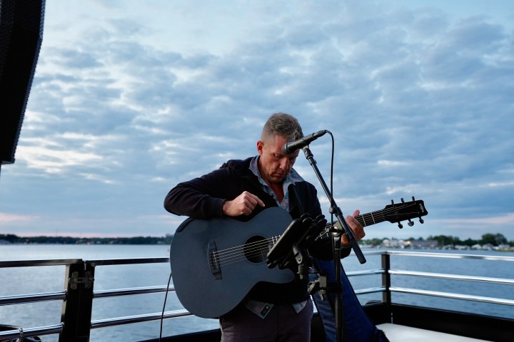 Man playing guitar on a boat with a cloudy sky and water in the background.