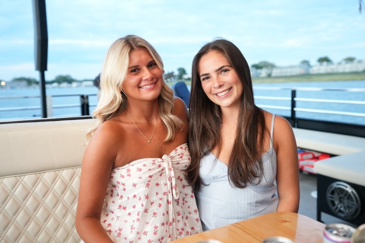 Two women smiling at an outdoor table with a water view backdrop.