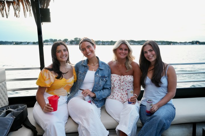 Four women sitting on a boat, holding drinks, with a water view in the background.