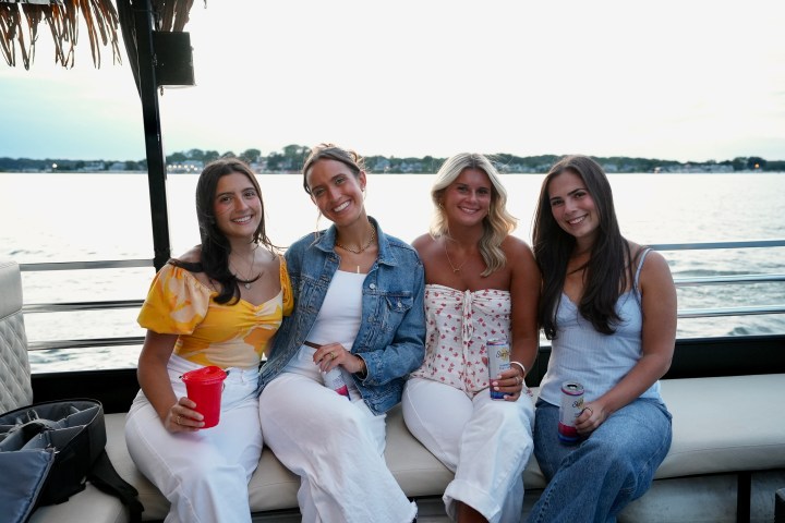 Four women sitting on a boat with drinks, smiling at the camera.