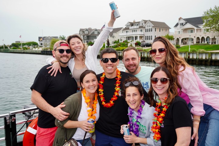 Group of people smiling with leis, one holding a drink, near waterfront homes.