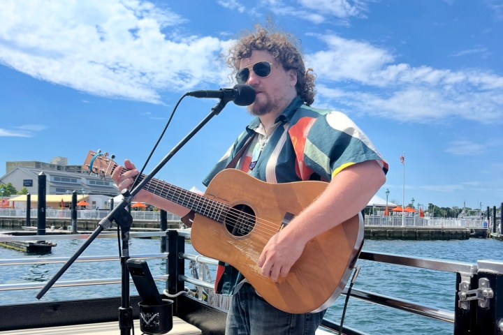Person with curly hair playing guitar and singing into a microphone on a boat with a waterfront background.