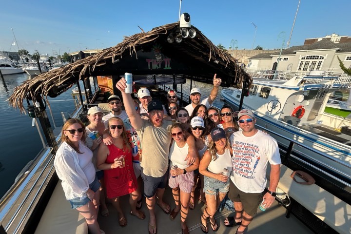 Group of people on a tiki-themed boat holding drinks, smiling, with marina in the background.