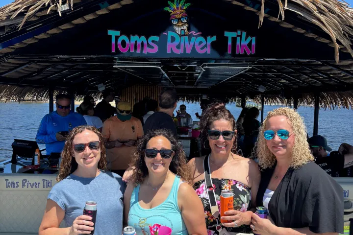 Four smiling women with drinks on a tiki boat under a blue sky.