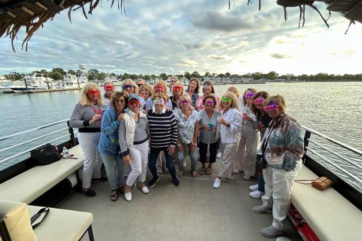 Group of women with colored glasses on a boat posing near a marina with cloudy sky.