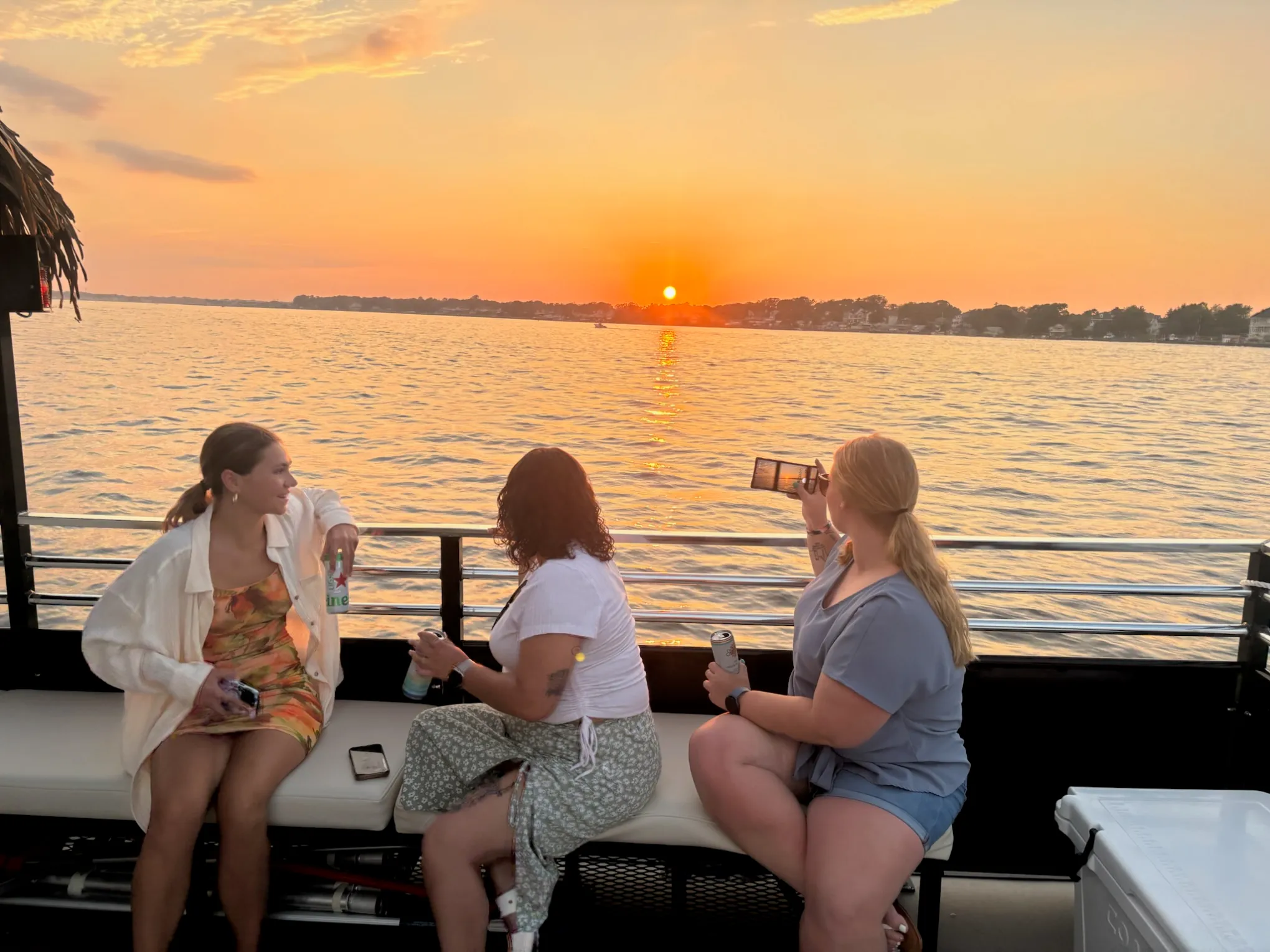 Three women on a boat at sunset, one taking a photo with a smartphone, others holding drinks.
