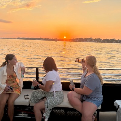 Three women on a boat at sunset, one taking a photo with a smartphone, others holding drinks.