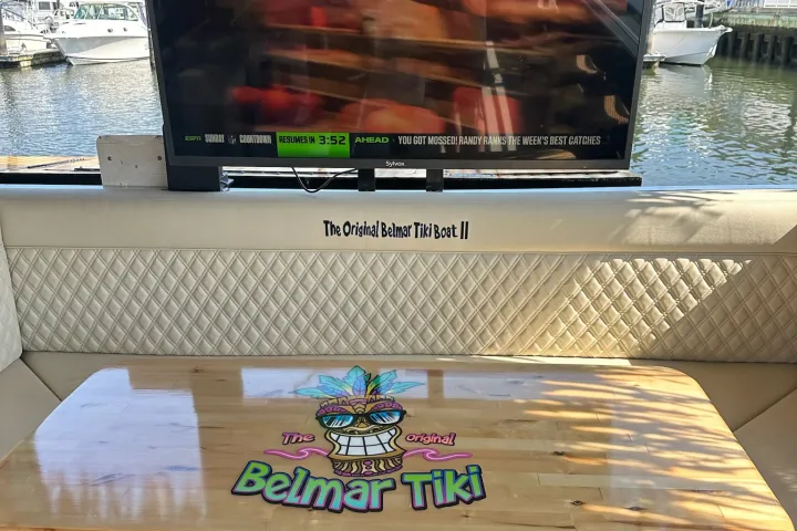 Outdoor seating with 'Belmar Tiki' logo on table, TV screen showing sports channel, boats in background.