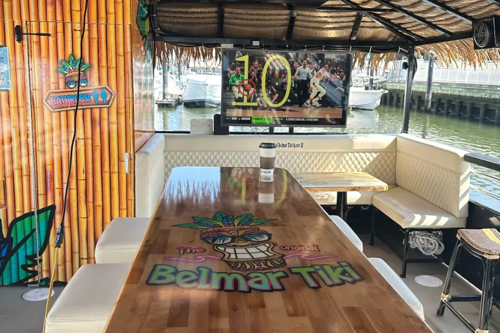 Boat interior with tiki bar, bamboo decor, TV, stools, and life vests overhead. 'Belmar Tiki' on bar top.