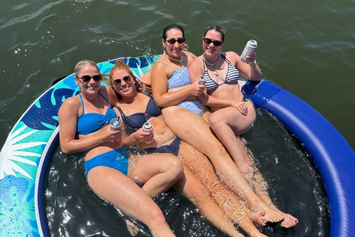 Four women on a blue inflatable ring in water, holding drinks and smiling.