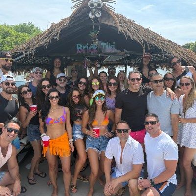 Group of people on a tiki boat with drinks, smiling, wearing summer clothes.