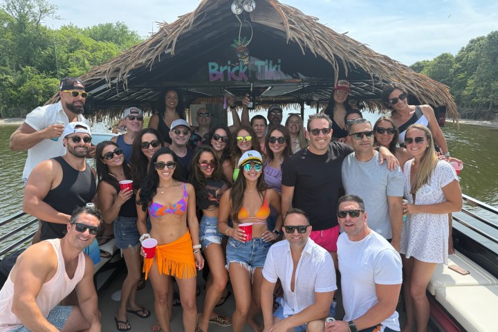 Group of people posing on a boat with tiki roof, wearing summer outfits and sunglasses.