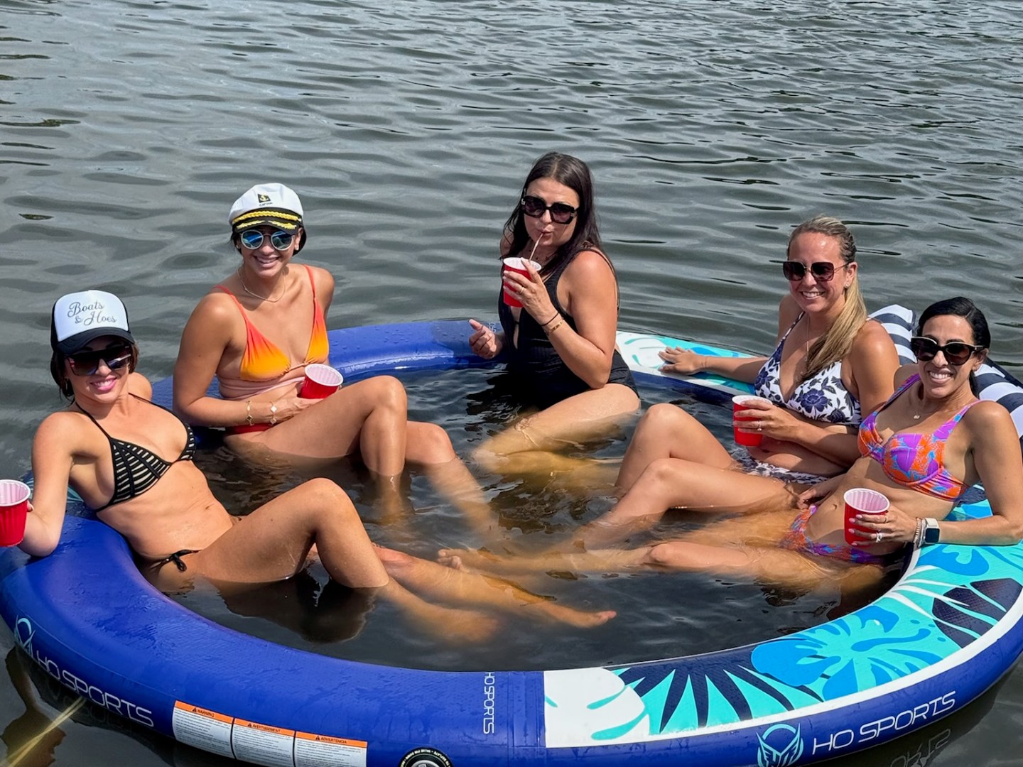 Five women relaxing on an inflatable float in the water, holding red cups and wearing swimsuits.