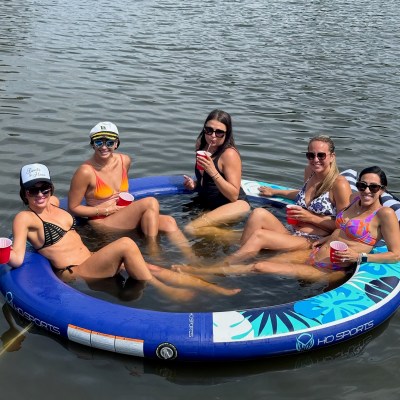 Five women relaxing on an inflatable float in the water, holding red cups and wearing swimsuits.