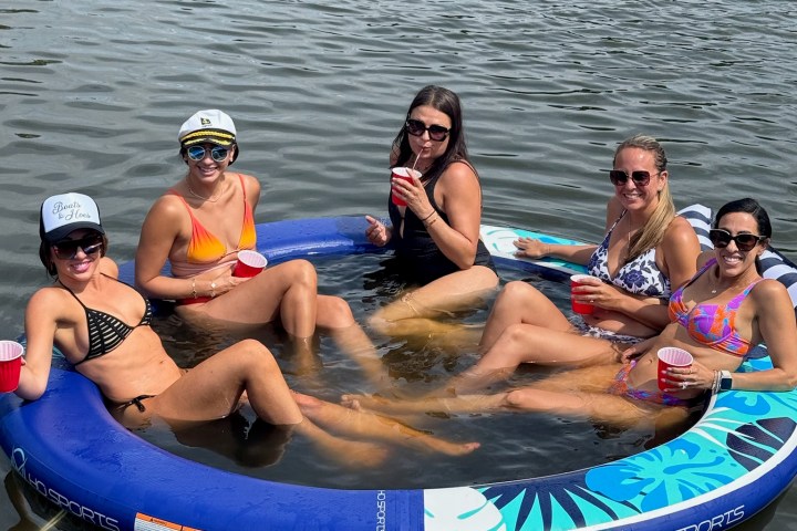 Five people relax on an inflatable lounger in a lake, holding red cups, wearing swimwear and sunglasses.