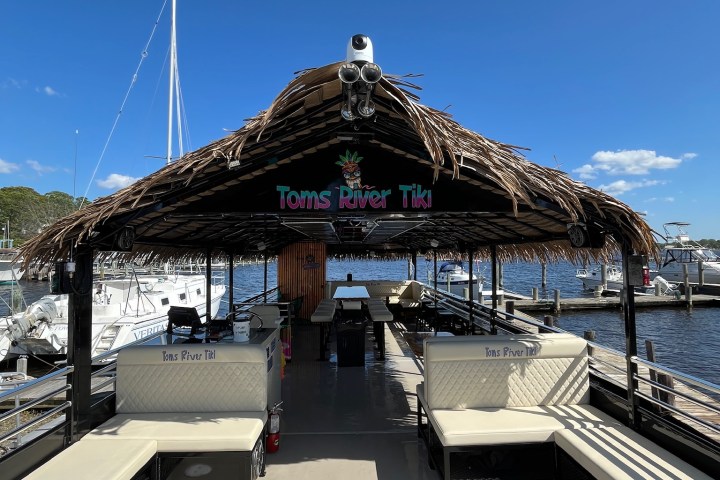 Tiki-themed boat docked at a marina, with benches and thatched roof, under a clear blue sky.