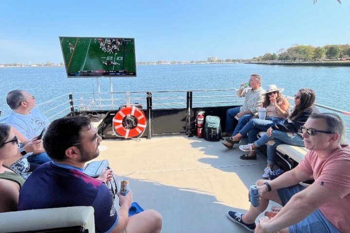 People on a boat watching a sports game on a TV under a clear blue sky.