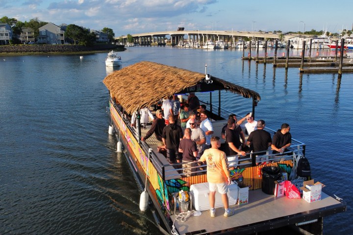 People enjoying a boat party on a river, with a bridge and marina in the background.