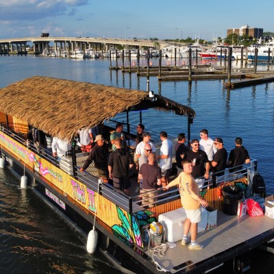 Group of people on a tiki boat with a thatched roof, docked near a marina on a sunny day.