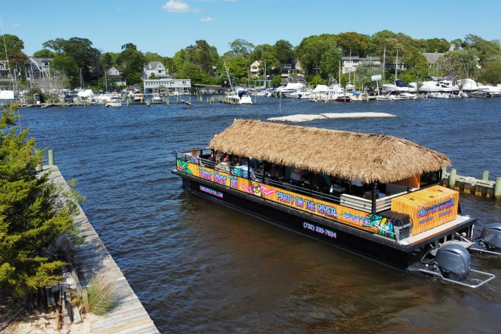 Tiki boat with thatched roof cruising on a river near a marina.