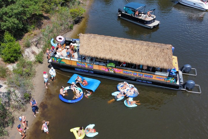 People relaxing on a river with boats and inflatables near a docked party boat with a thatched roof.