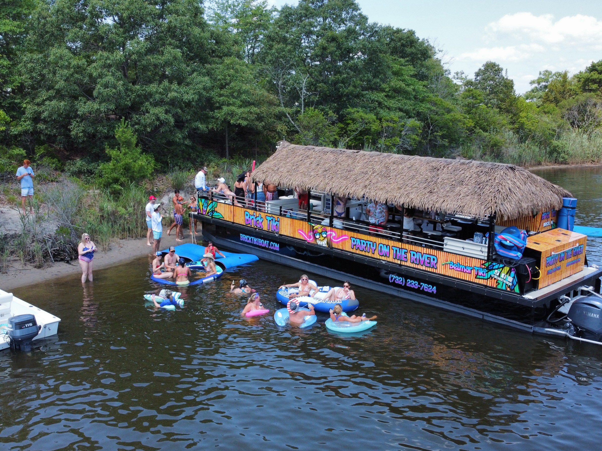People on a pontoon boat and others floating in the water near a wooded shoreline.