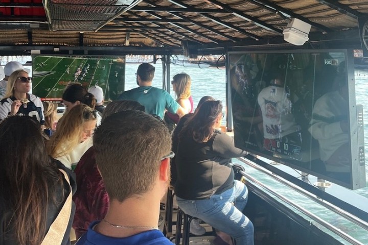 People on a boat watching sports on TV screens with water in the background.