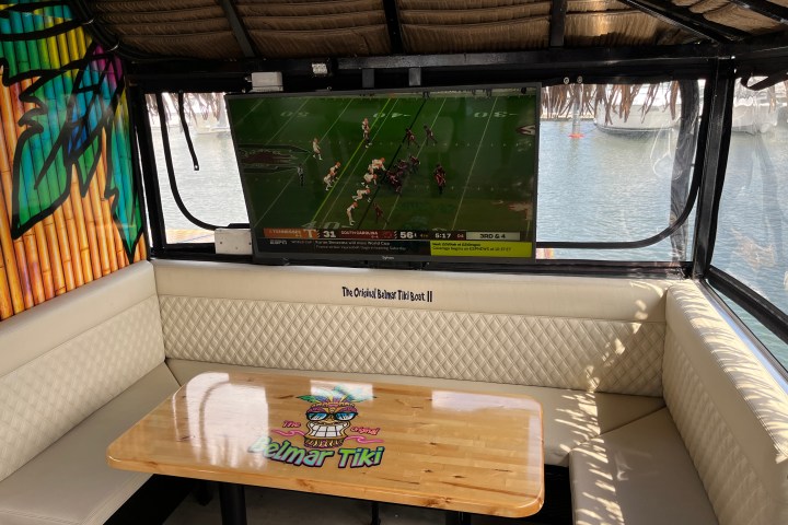 Boat interior with a TV showing a football game, a table with Tiki branding, and water view outside.