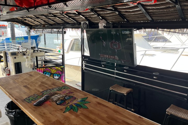 Outdoor bar on a boat with a TV showing a football game and a colorful sign on the wooden counter.