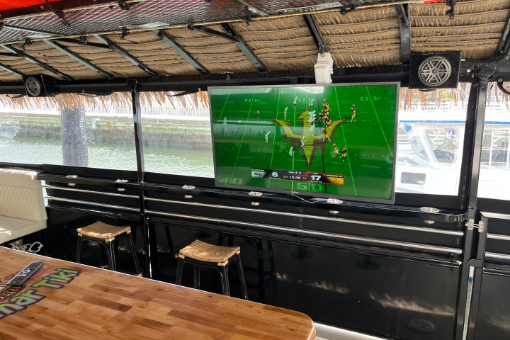 TV showing a football game in a room with wooden table and stools, thatched roof, and lake view.