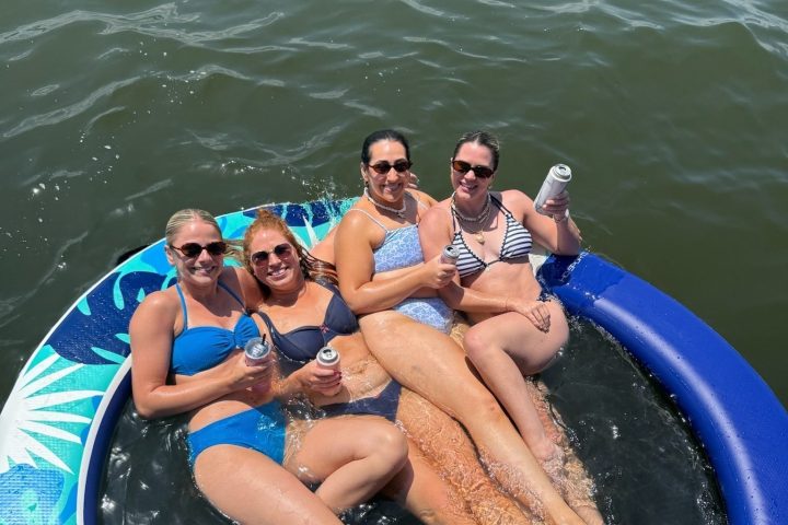 Four women in swimsuits relaxing on a large blue inflatable ring in the water, holding beverage cans.