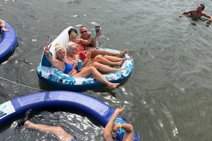 Group of people relaxing on inflatables in a lake, holding drinks, with boats and a swimmer nearby.