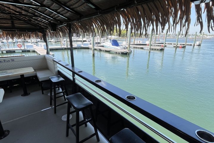 Outdoor bar with stools by a calm marina under a thatched roof.