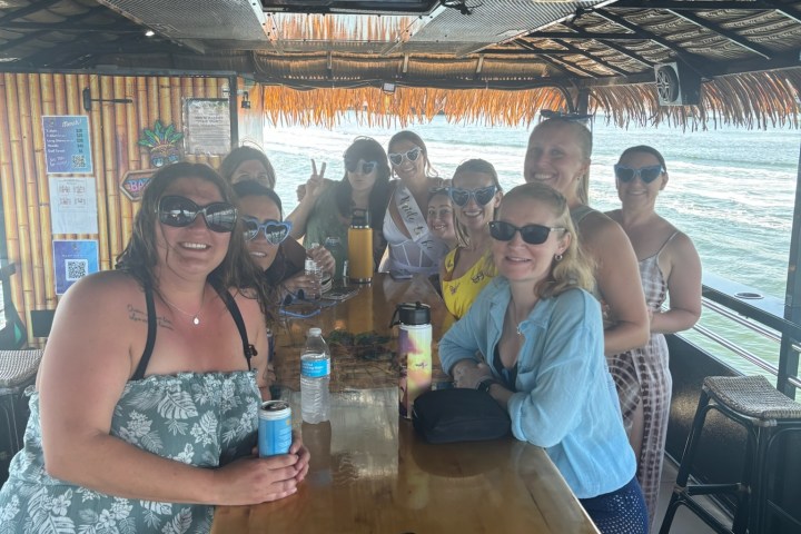 Group of people sitting at a wooden bar under a thatched roof, smiling at the camera.